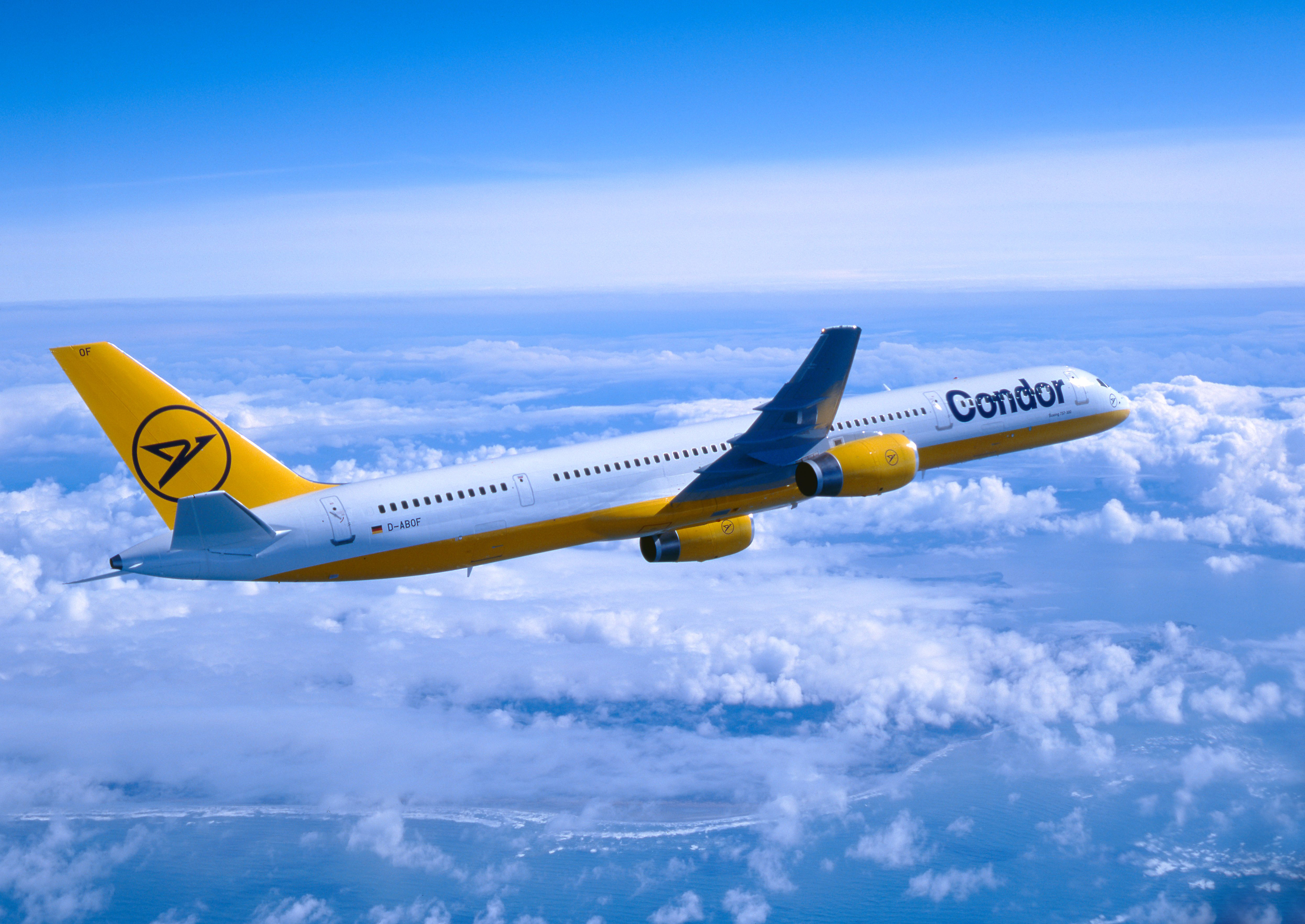 Historical image of a Condor aircraft in yellow and white livery flying over closed cloud cover. The aircraft is shown from the side against a blue sky.