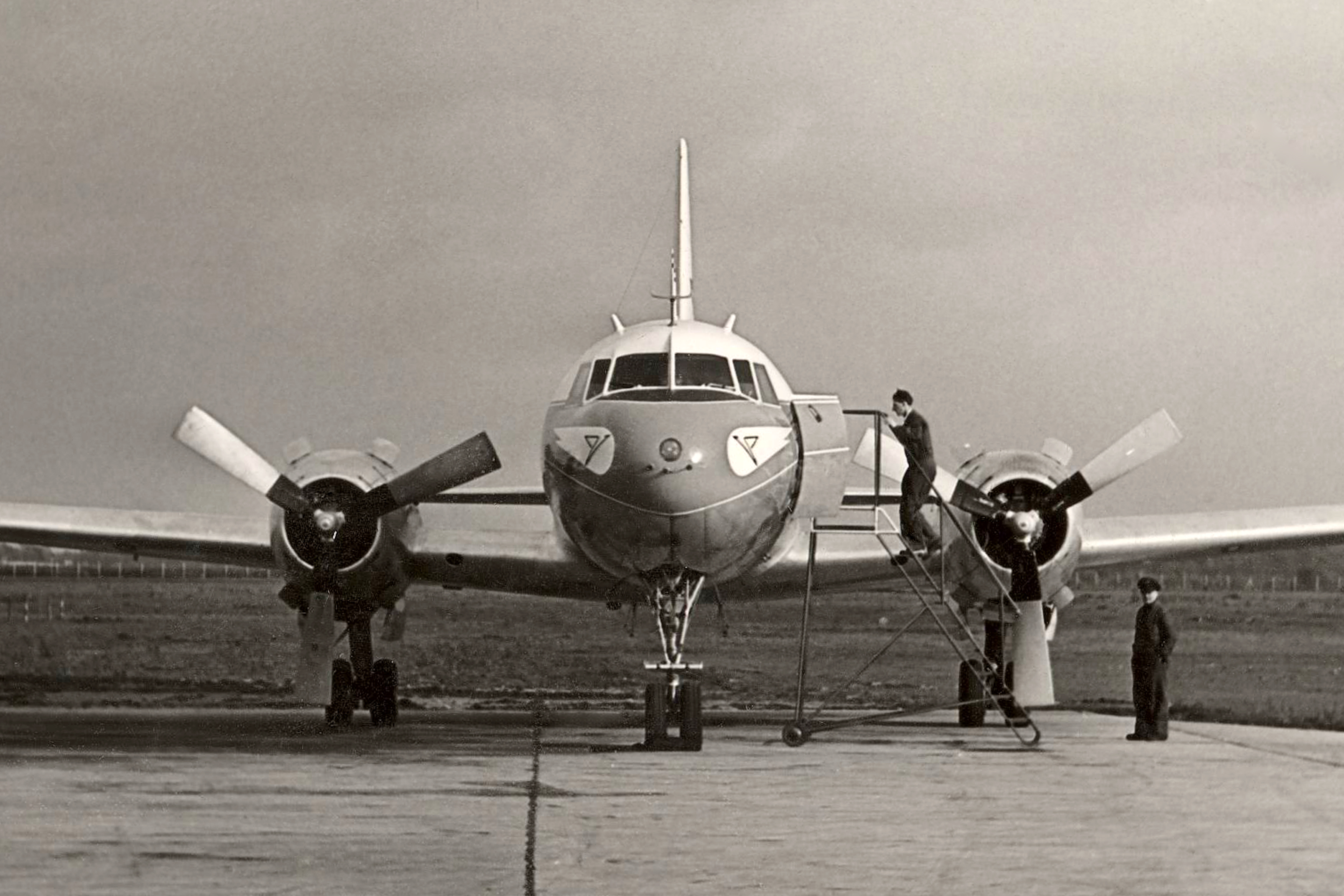 The picture shows boarding in 1958 via a mobile staircase into a Convair CV-240. The historic propeller-powered passenger plane is seen from the front on the apron of an airport. One person stands on the stairs at the open plane door while another person stands next to the plane. The photograph is in black and white.