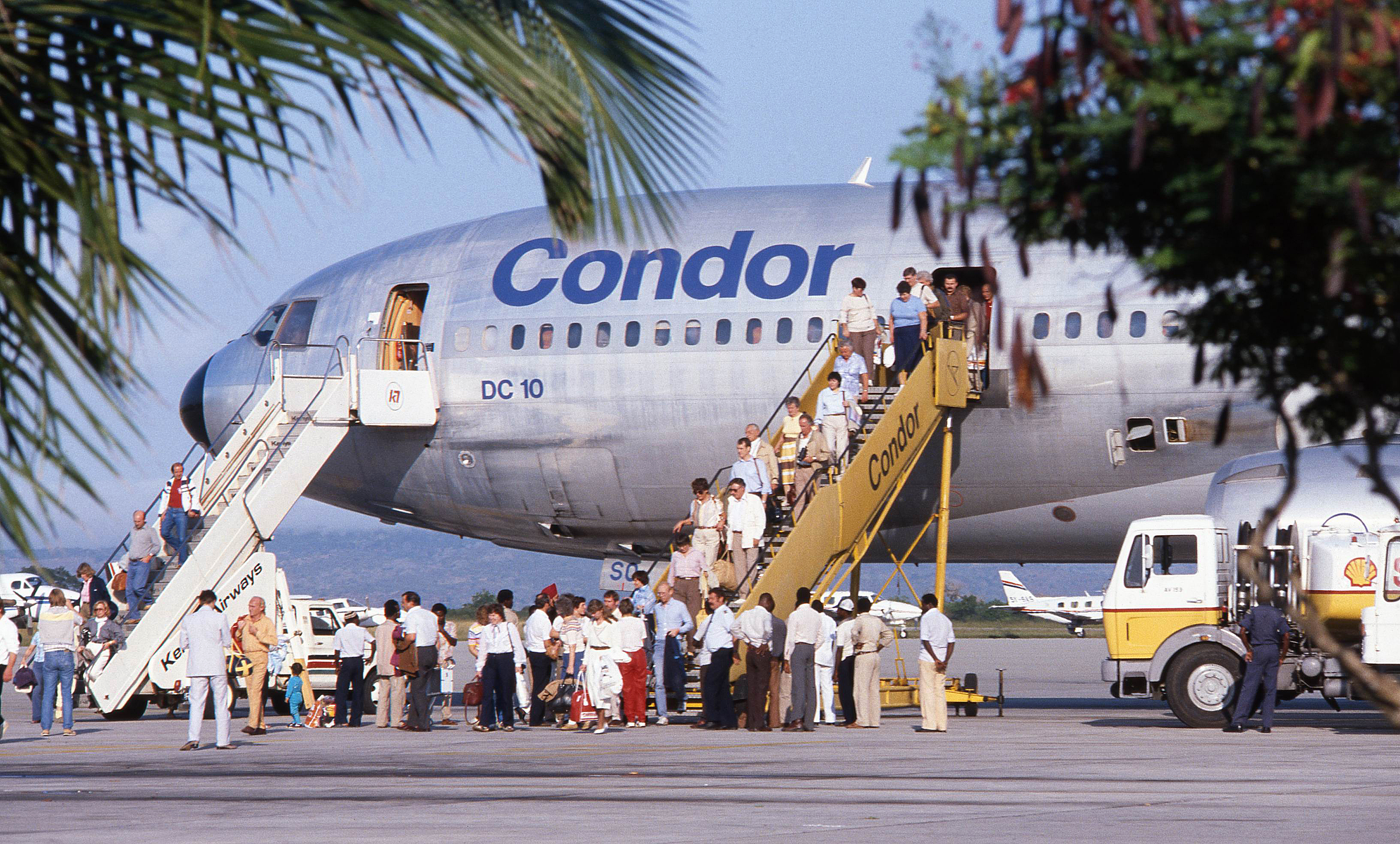 The picture shows passengers boarding and disembarking from a vintage passenger aircraft via two mobile staircases. There are several people on the steps and on the apron in front of the airplane. The aircraft is shown in profile. Airport vehicles can be seen in the foreground, while palm fronds frame the picture.